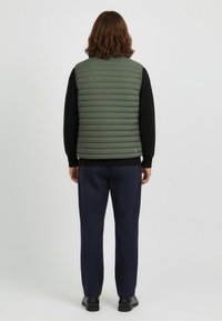 Green puffy vest with vertical quilting, worn over a black ribbed sweater, paired with navy trousers and black shoes.