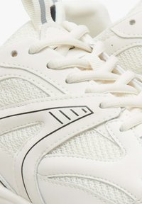Białe buty sportowe z siateczkowej tkaniny i skóry syntetycznej. Sznurowadła i szary pasek akcentują gładki i fakturowany design.