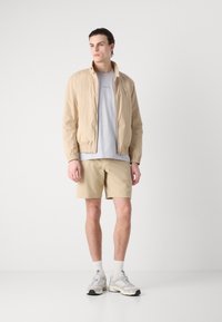 Chaqueta de nailon beige claro con cierre de cremallera, combinada con una camiseta gris claro y pantalones cortos beige texturizados, complementada con zapatillas blancas y grises.