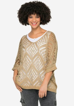 Pull en crochet beige clair avec un motif en losanges et des manches larges, superposé à une chemise blanche. Texture libre et ample.