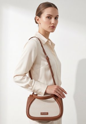 Jeune femme aux cheveux plaqués porte un chemisier crème et un sac à main beige et marron Diana&Co Firenze à l'épaule.