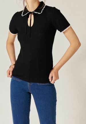 Kvinde iført en sort kortærmet top med hvid blonderkant på krave og ærmer, kombineret med blå jeans.