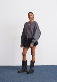 Mujer con trenzas largas que lleva un sudadera gris de Nike de gran tamaño, una falda negra con pliegues y botas altas negras, de pie contra una pared blanca sobre una alfombra azul.