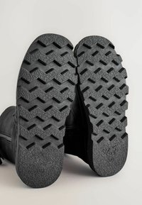 Svarta stövlar med tjocka, mönstrade gummisulor vända uppåt, som visar djup zickzack och texturerat mönster för grepp.
