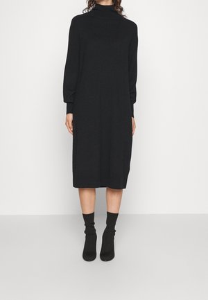 Femme portant une longue robe noire à col roulé avec des manches bouffantes, associée à des bottines noires mi-mollet, se tenant devant un fond uni.