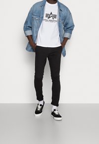 Chaqueta de mezclilla, camiseta blanca con logo negro, pantalones ajustados negros y zapatillas negras con suelas blancas, contra un fondo liso.