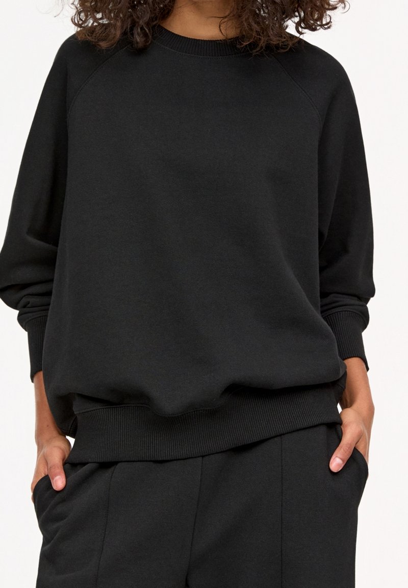 Personne portant un sweat-shirt noir ample et un pantalon noir assorti, mains dans les poches, sur un fond clair uni.