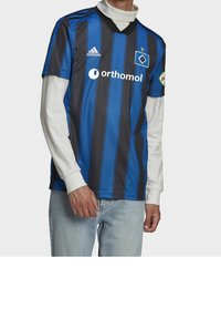 Modrý a černý pruhovaný fotbalový dres s výstřihem do V, krátkými rukávy a logem Adidas; kombinovaný s bílou košilí s dlouhými rukávy pod ním.