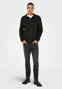Donkergroene kabelgebreide trui met een kraag, voorzien van een gewatteerd patroon. Gecombineerd met zwarte slim-fit jeans en zwarte laarzen.
