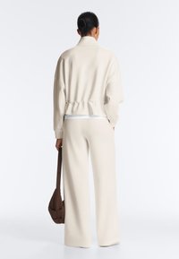 Personne aux cheveux courts et foncés portant un pull à col roulé crème et un pantalon à jambes larges, tenant un sac à bandoulière marron, de dos.
