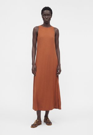 Kvinde stående iført en ærmeløs, rustfarvet midi-kjole med sideslids og brune flade sandaler mod en ensfarvet hvid baggrund.