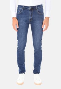 Uomo che indossa jeans slim-fit blu e sneakers bianche, in piedi con le braccia rilassate ai lati, su uno sfondo bianco semplice.