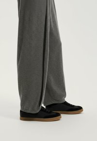 Szare szerokie spodnie z miękkiego materiału, zestawione z czarnymi trampkami o gładkiej cholewce i teksturowanej, brązowej gumowej podeszwie.