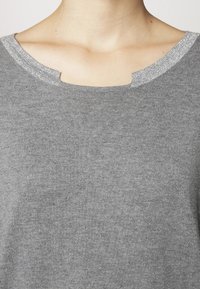 Grauer Pullover aus strukturiertem Stoff, mit rundem Ausschnitt und einem metallischen Akzent am Kragen. Minimalistisches Design, keine Muster.
