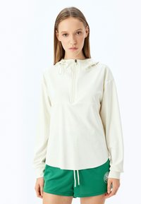 Top de manga larga crema claro con media cremallera, capucha ajustable y corte relajado, combinado con pantalones cortos deportivos verdes que presentan un logo.
