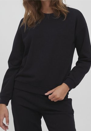 Kvinde iført ensfarvet sort langærmet sweatshirt og matchende sorte bukser, med løst hår, stående mod en lysegrå baggrund.
