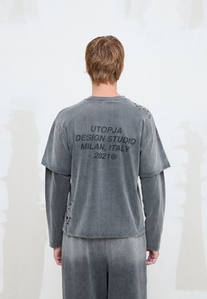 Persona con cabello corto castaño claro lleva una camiseta gris descolorida con varias capas, de manga larga y corta, con el texto "UTOPJA DESIGN STUDIO MILAN, ITALY 2021" en la espalda.