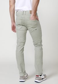 Jeans slim-fit verde claro vistos desde atrás, hechos de denim con una textura suave. Cuentan con dos bolsillos traseros y una etiqueta de cuero marrón.