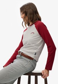 Kobieta siedząca bokiem na drewnianym stołku, ubrana w szary i czerwony długi rękaw, w paskowane spodnie oraz brązowy pleciony pasek, patrząca w dół.