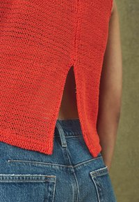 Rød strikket top med åben rygdetalje, parret med højtaljede blå denim jeans. Teksturen er løs og mesh-lignende.