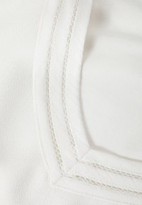 Tissu blanc avec une texture lisse, présentant un design de col en couches et des coutures décoratives le long des bords.