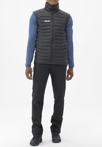 Gilet rembourré noir avec des boudins horizontaux, col haut et fermeture éclair à l'avant, associé à un haut ajusté à manches longues bleu et un pantalon foncé.