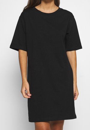 Kobieta ubrana w prostą czarną sukienkę T-shirt z krótkim rękawem sięgającą kolan, stojąca na białym tle.