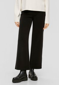 Zwarte wijde broek van zacht materiaal, met een gladde textuur en een losse pasvorm. G gedragen met dikke zwarte enkellaarzen.