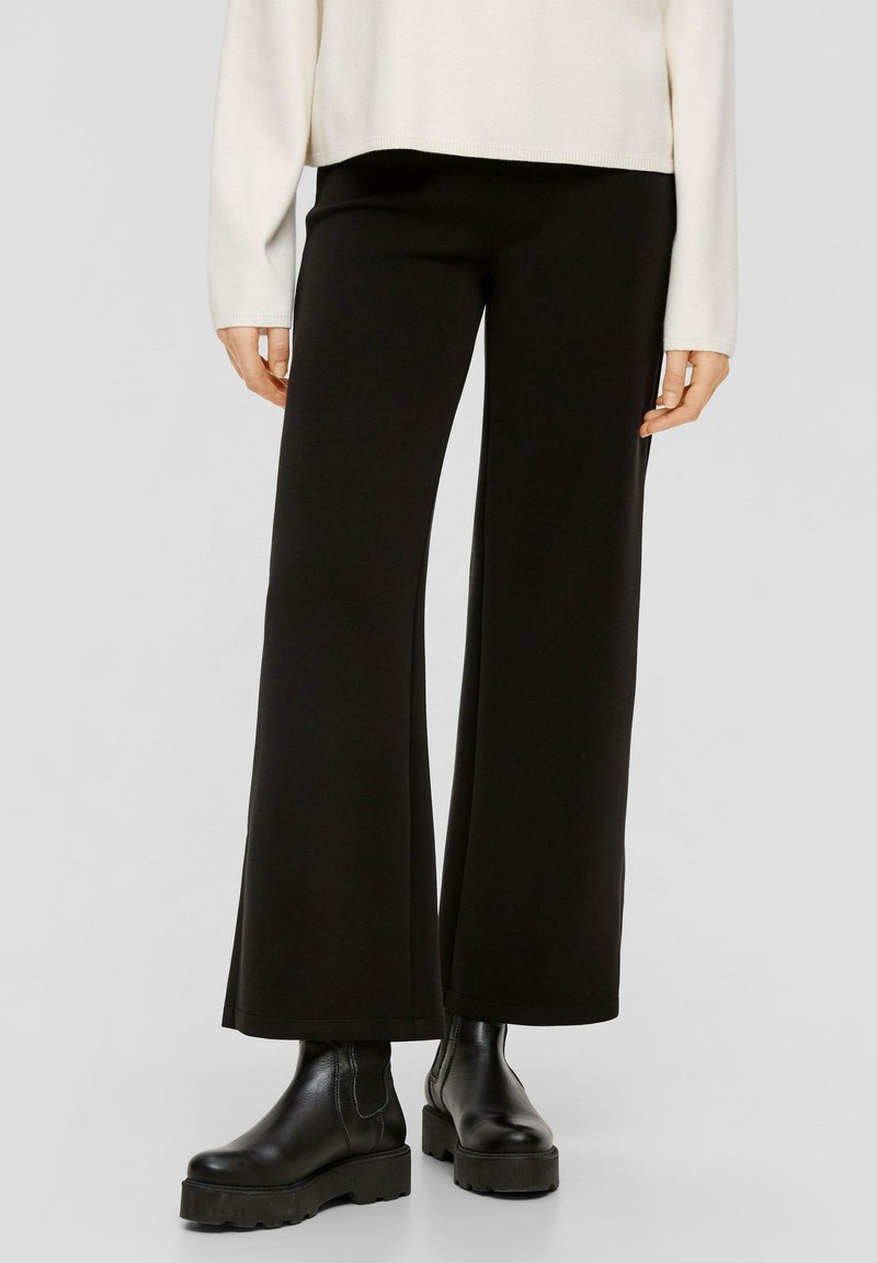 Zwarte wijde broek van zacht materiaal, met een gladde textuur en een losse pasvorm. G gedragen met dikke zwarte enkellaarzen.