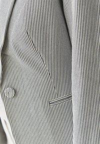 Pruhovaný blazer s vertikálními námořnickým a bílými pruhy, klopovým límcem, jedním knoflíkem a dekoračním detailem kapsy.