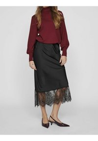 Jupe en satin noir avec ourlet en dentelle, associée à un haut bordeaux à manches longues. Portée avec des escarpins sombres à bout pointu. Texture lisse et design élégant.