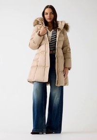 Beżowa kurtka puchowa z kapturem wykończonym futerkiem, zapinana na zamek, z pikowaną fakturą, w połączeniu z paskowaną koszulą i rozszerzanymi dżinsami.