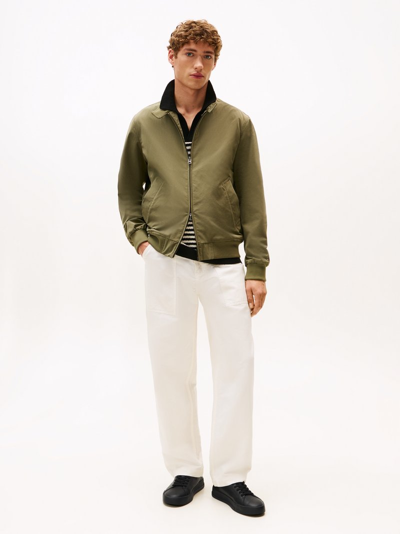 Casaco bomber verde-oliva com fecho de zip, gola preta, combinado com calças brancas e sapatilhas pretas. Textura suave, design casual.
