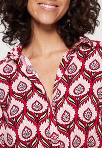 Czerwona koszula w kwiaty z różowymi kwiatami i zielonymi liśćmi, z kołnierzykiem zapinanym na guziki oraz gładką fakturą materiału z detalicznymi wzorami kwiatowymi.