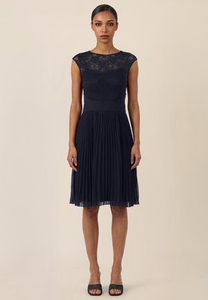 Frau steht und trägt ein marineblaues Kleid mit Spitzenoberteil und plissiertem Rock, kombiniert mit schwarzen offenen Pumps, neutraler Hintergrund.