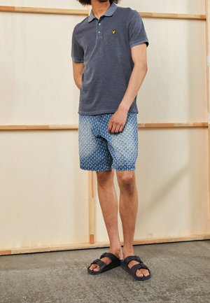 Mężczyzna w ciemnoszarej koszulce polo, wzorzystych niebieskich szortach i czarnych sandałach na podwójny pasek stoi w pomieszczeniu na tle drewnianym i beżowym.