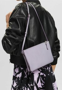 Světle fialová kožená kabelka přes rameno se zipovým zapínáním, štíhlým tvarem, nastavitelným popruhem a minimálními kovovými detaily. Nositelná přes černou bundu.