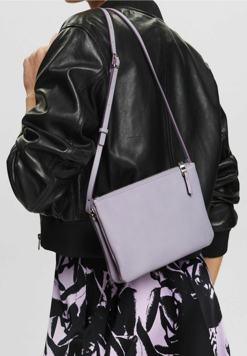 Světle fialová kožená kabelka přes rameno se zipovým zapínáním, štíhlým tvarem, nastavitelným popruhem a minimálními kovovými detaily. Nositelná přes černou bundu.
