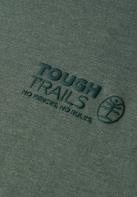 Zelené bavlněné tričko s texturovaným potiskem nápisu "TOUGH TRAILS" a kruhovým logem stromu. Materiál má jemnou a hladkou texturu.