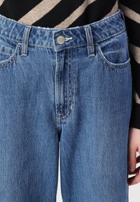 Zilas džinsus ar augstu jostasvietu, priekšējiem kabatām un neapstrādātu apakšmali. Audumam ir gaiša mazgāšana un smalka tekstūra ar kontrastējošu vītni.