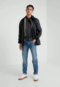 Jovem vestido com uma jaqueta preta sobre uma camisa polo preta, jeans azuis e ténis brancos, de pé com uma mão no bolso contra um fundo simples.
