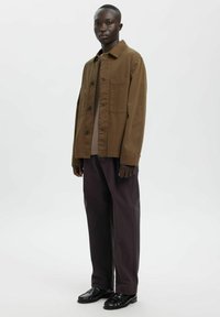 Brązowa bawełniana kurtka koszulowa z kołnierzem i przednią kieszenią, zestawiona z ciemnofioletowymi spodniami i czarnymi mokasynami. Prosty, uporządkowany fason.