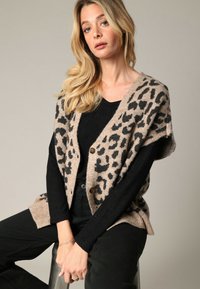 Cardigan beige avec imprimé léopard noir, doté d'un col en V, de grands boutons et d'une coupe ample, associé à un haut noir côtelé et un pantalon noir.