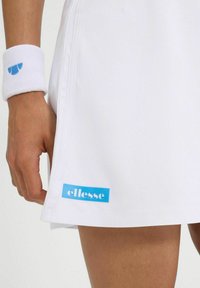 Weißer Tennisrock mit glatter Textur, mit einem blauen "ellesse"-Logo am Saum. Begleitet von einem weißen Handgelenkband mit blauem Logo.