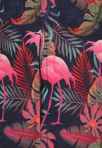 Shorts med en mørk marineblå baggrund, der har et mønster af en lys pink flamingo og tropiske blade. Stoffet har en glat, let tekstur.