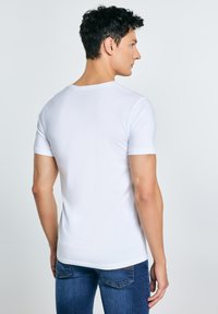 Hombre de espaldas con camiseta blanca de manga corta lisa y jeans azules, frente a un fondo claro y uniforme.