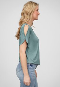 Blouse turquoise à manches courtes avec des épaules dénudées, coupe décontractée et tissu doux. Portée avec un jean bleu clair.