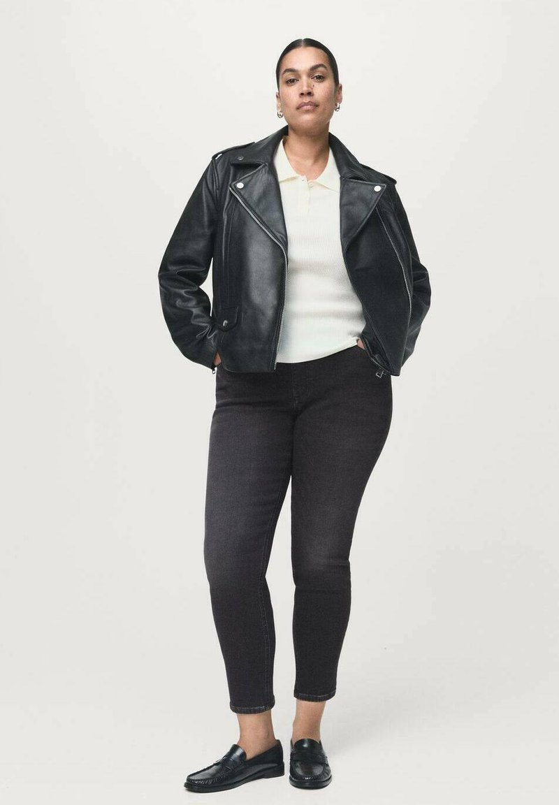 Una mujer se mantiene erguida y confiada, vistiendo una chaqueta de cuero negra, una camiseta polo blanca, unos vaqueros negros ajustados y unos mocasines negros, todo ello sobre un fondo liso.