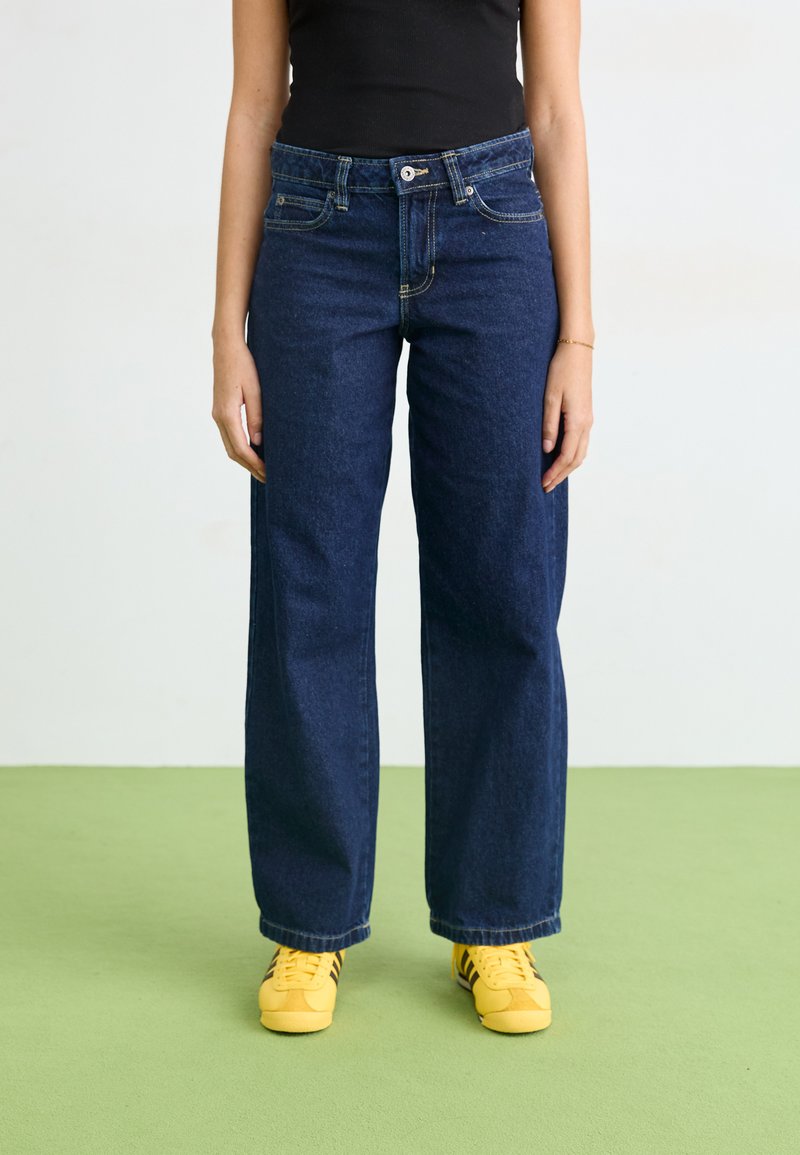 Jeans a gamba larga di un blu scuro con vita alta, caratterizzati da un'ormai classica combinazione di cinque tasche e dettagli di cucitura discreti. Abbinati a sneakers gialle.