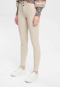 Pantalones vaqueros ajustados beige con una cintura alta, que presentan dos botones frontales y una textura suave. Combinados con zapatillas blancas con cordones.